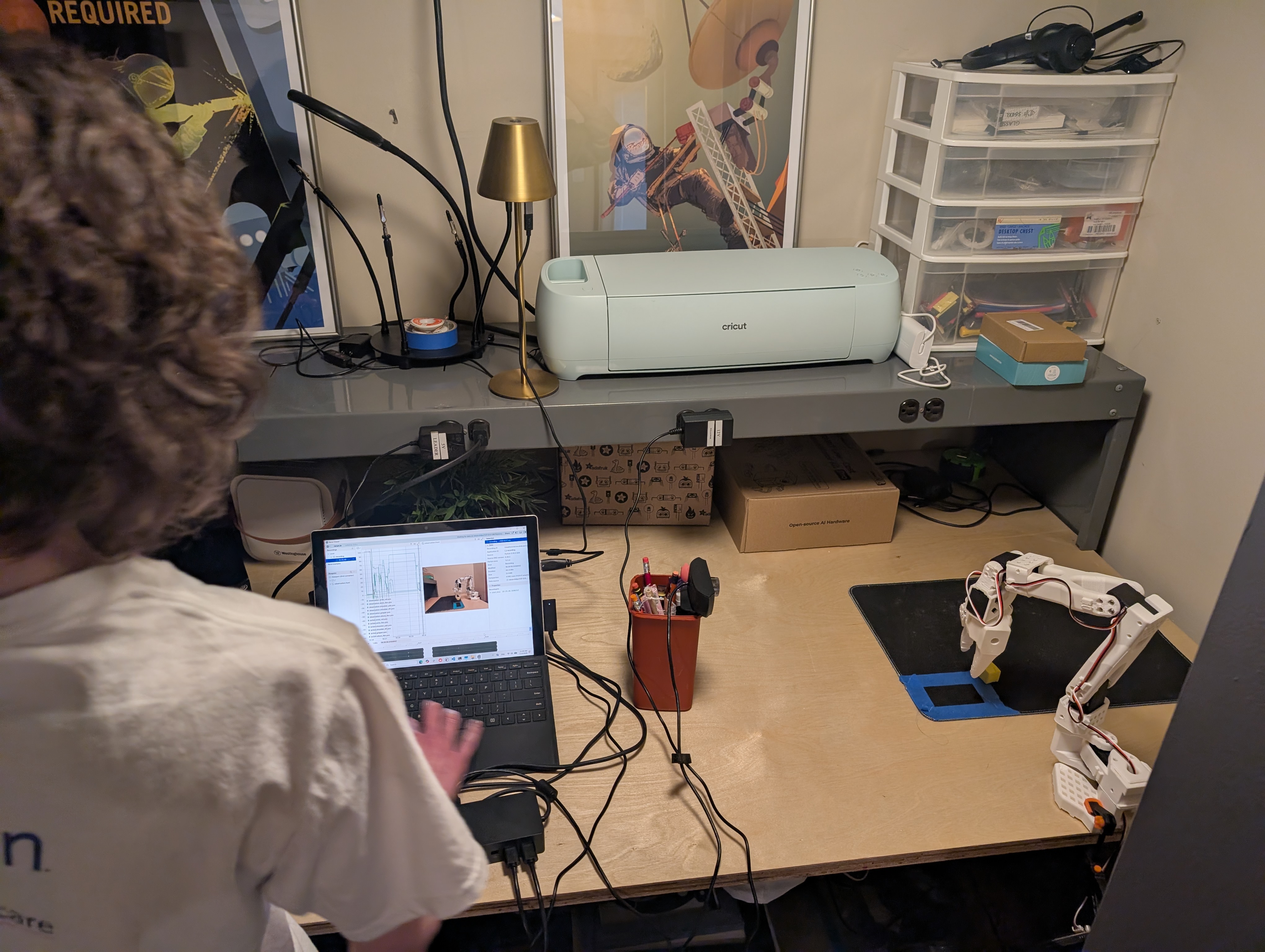 Home workbench with the SO-101 follower arm on a tape-marked mat, a recording laptop, and maker gear on the shelf above